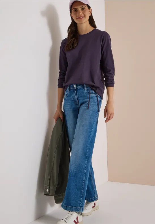 Une femme appuyée contre un mur, porte un top violet à manches longues, un jean ample, des baskets blanches et une casquette rose.