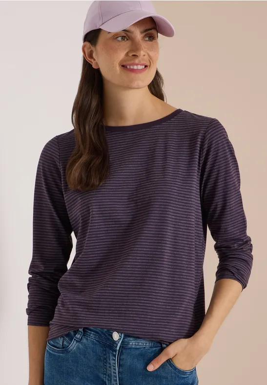 Femme souriante en casquette violette et t-shirt à rayures, main dans la poche, regard de côté.