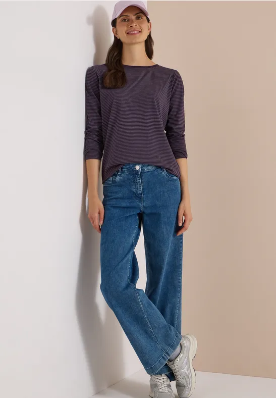 Jeune femme appuyée à un mur clair, portant un tee-shirt violet à manches 3/4, jean bleu, baskets blanches et casquette rose.