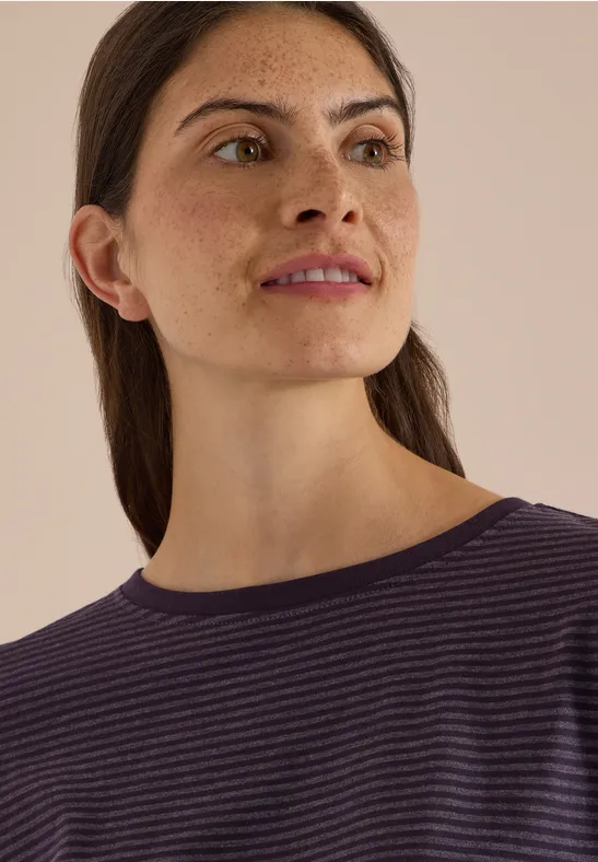 Portrait d'une femme aux taches de rousseur, cheveux attachés, en t-shirt violet à fines rayures, regard de côté.