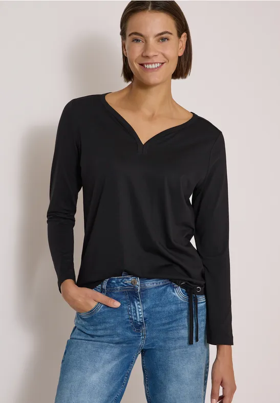 Eine Frau mit schwarzem Langarmshirt und blauen Jeans, lächelnd vor neutralem Hintergrund.