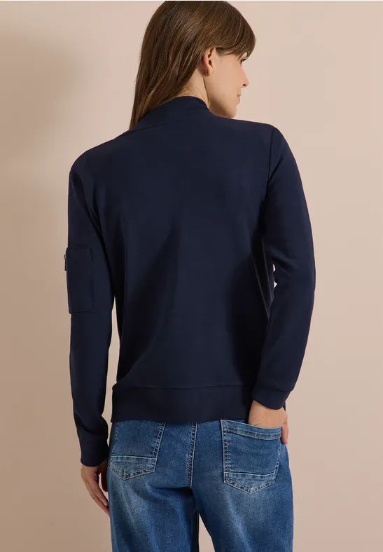 Frau von hinten in dunkelblauer Sweatjacke und Jeans vor beiger Wand.