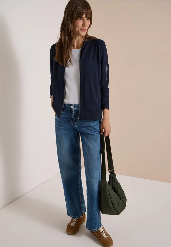 Frau in weißem T-Shirt, dunkelblauer Strickjacke, weiten Jeans, braunen Sneakern, grünem Schulterbeutel.