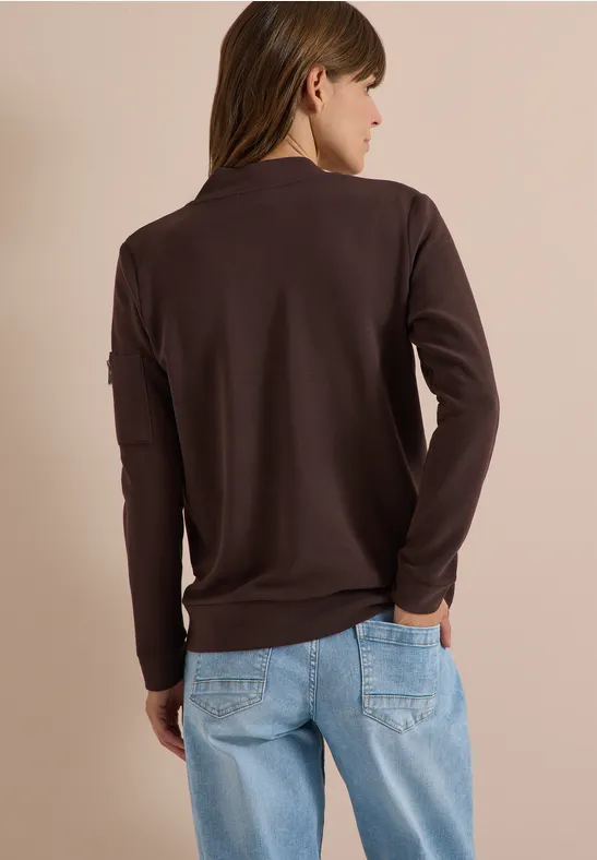 Eine Frau von hinten mit braunem Langarmshirt und hellblauen Jeans vor beigem Hintergrund.