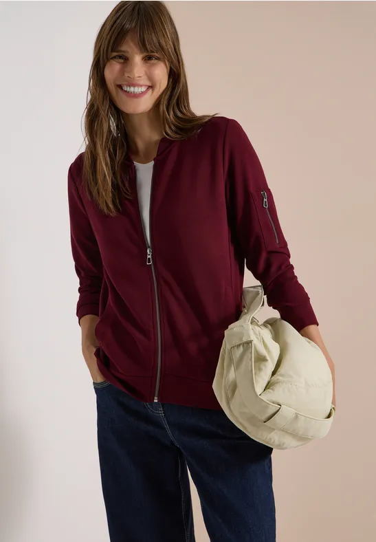 Fröhliche Frau in burgunderroter Jacke, weißem Shirt und Jeans hält eine beige Tasche.