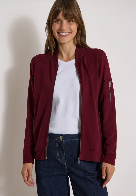 Eine lächelnde Frau trägt eine bordeauxrote Jacke, weißes T-Shirt und blaue Jeans vor hellem Hintergrund.
