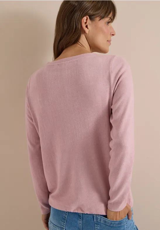 Une femme vue de dos porte un pull rose à manches longues et un jean devant un mur aux tons neutres.