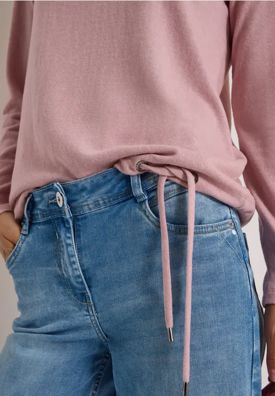 Pull à manches longues rose avec cordon à la taille, glissé dans un jean bleu, mains dans les poches.
