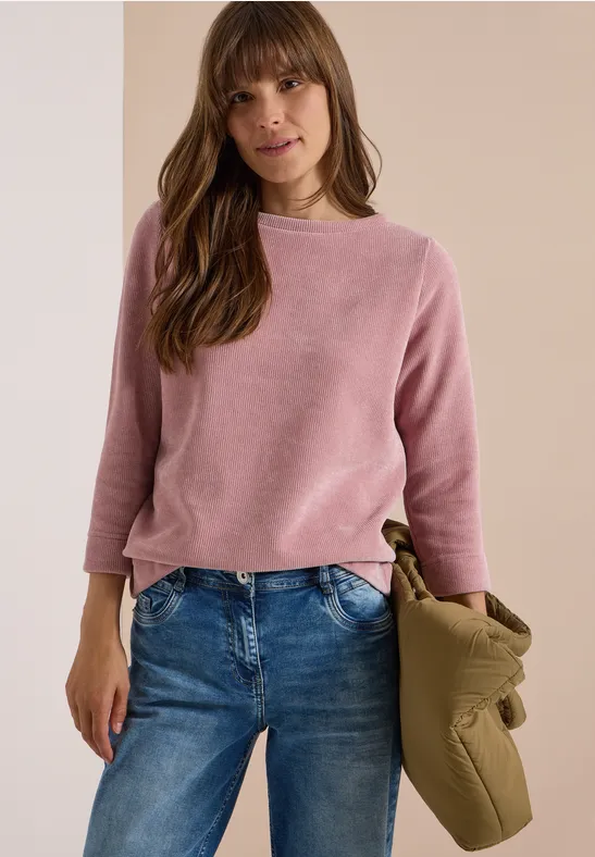 Femme en pull rose et jeans, tenant une veste kaki, posant devant un fond neutre.