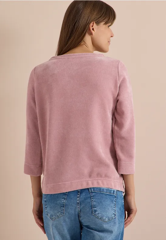 Personne de dos portant un pull douillet rose à manches 3/4 et un jean bleu sur fond beige.