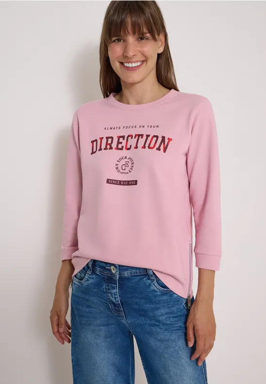 Femme souriante portant un sweat rose avec l’inscription « DIRECTION » et un jean bleu.