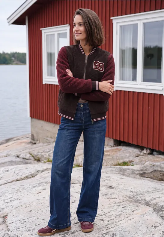 Frau in Jeans und Collegejacke steht vor einem roten Haus am See auf Felsen.