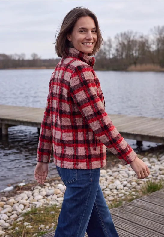 Une jeune femme en veste à carreaux sourit en regardant par-dessus son épaule au bord d’un lac.