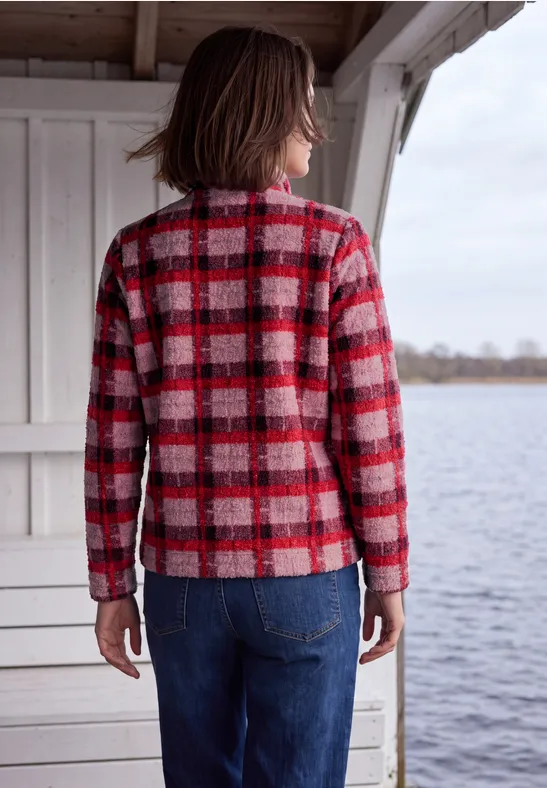 Personne portant un pull à carreaux rouge et un jean bleu, debout sous un abri en bois face à l'eau.
