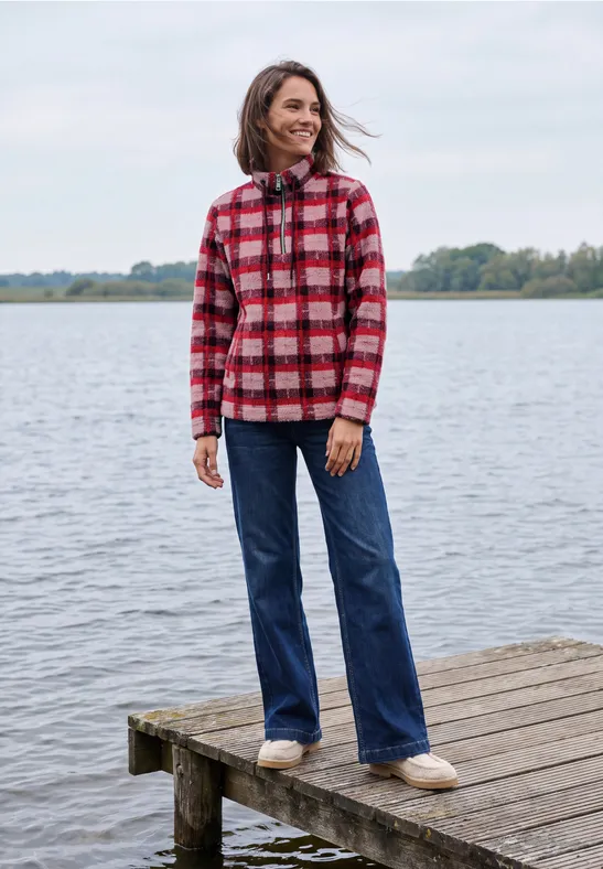Une femme en chemise à carreaux rouge sourit sur une jetée en bois au bord d'un lac paisible.