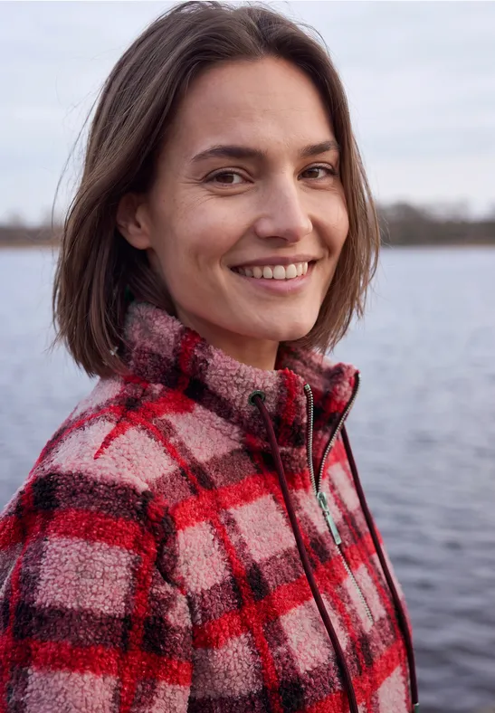 Une femme aux cheveux bruns sourit, portant une veste à carreaux rouge et noir près d'un lac.