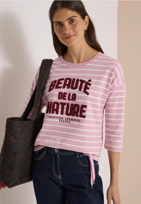 Femme portant un t-shirt rose à rayures 'Beauté de la nature', jean et sac noir.