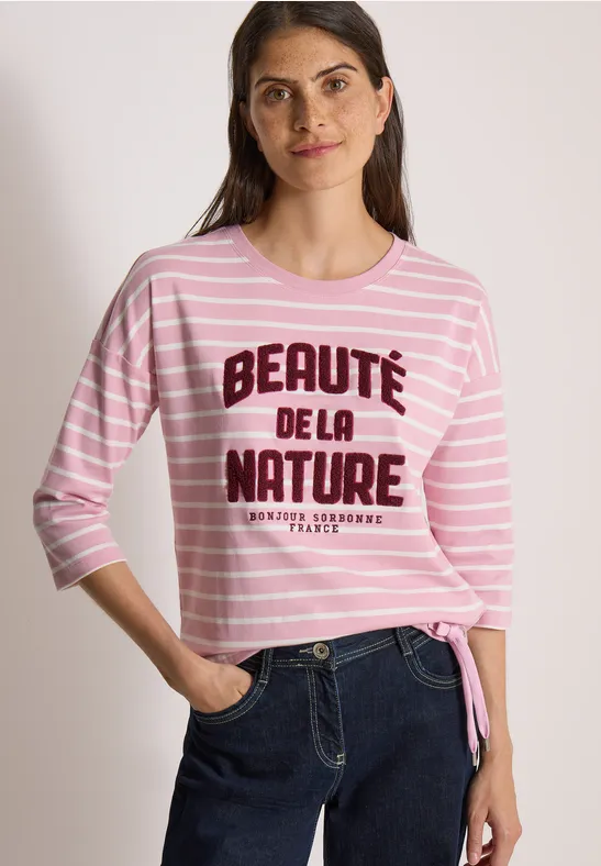 Une femme souriante porte un t-shirt rayé rose et blanc 'Beauté de la nature' et un jean.