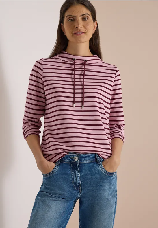Femme en sweat à capuche rose à rayures noires et jean bleu, mains dans les poches, fond neutre.