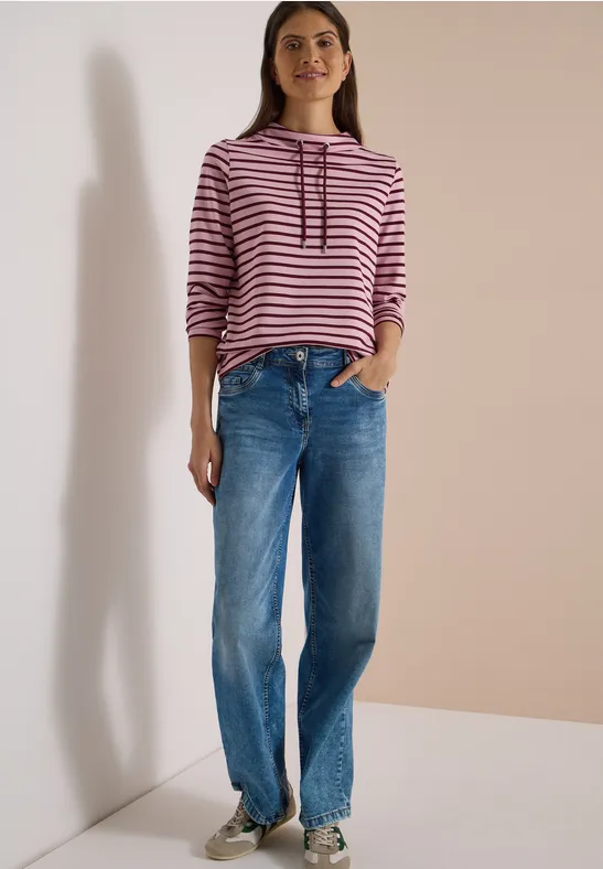 Femme en sweat à capuche rayé rose et blanc et jean bleu large, posant devant un fond neutre.