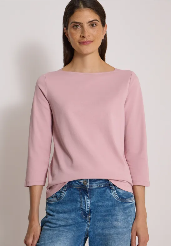 Femme portant un pull rose et un jean bleu devant un fond neutre.