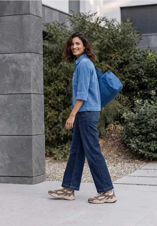 Eine junge Frau in Jeans und Sneakers mit blauem Rucksack geht entspannt auf dem Bürgersteig.
