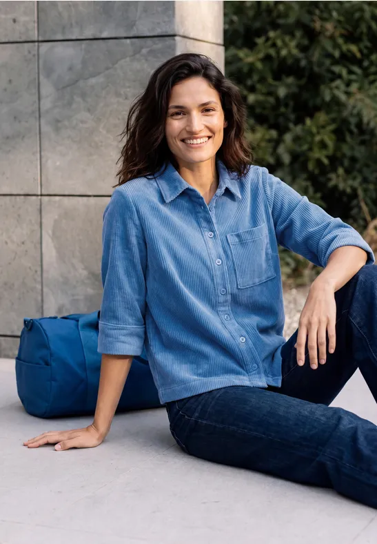 Eine Frau sitzt entspannt vor einer Mauer, trägt Jeanshemd und lächelt, eine blaue Tasche liegt neben ihr.
