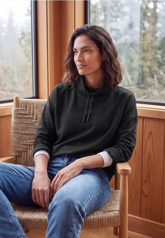 Femme en pull noir assise dans un fauteuil en bois, le regard tourné vers l'extérieur.