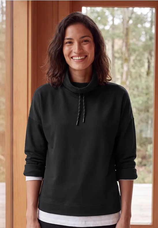 Une femme souriante en sweat noir et T-shirt blanc, debout devant une fenêtre en bois.