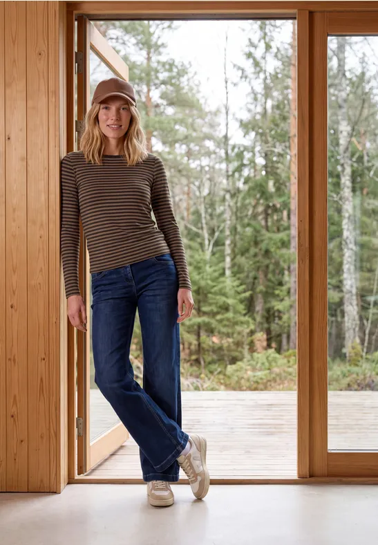Une femme en t-shirt à rayures, jean et bonnet se tient détendue dans une pièce en bois devant de grandes fenêtres.