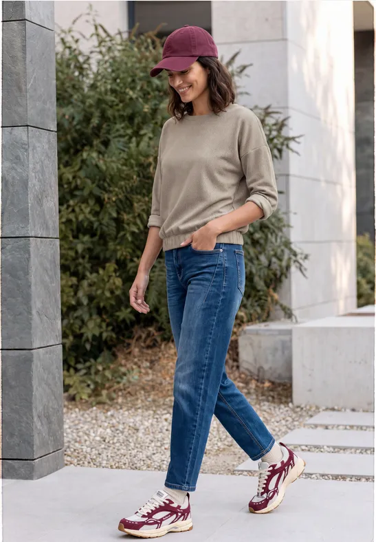 Junge Frau mit Jeans, Pullover, roter Kappe und Turnschuhen geht draußen spazieren und lächelt.