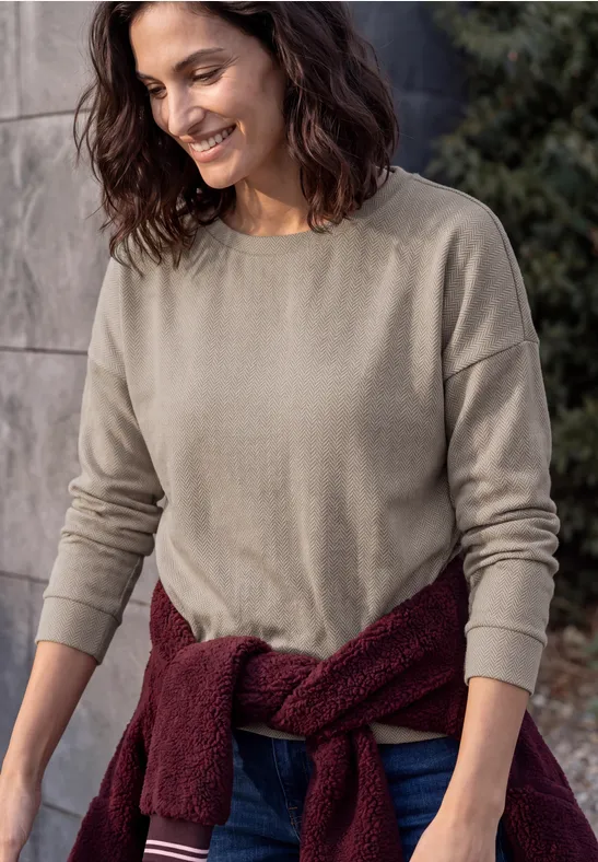 Eine lächelnde Frau in beige Langarmshirt, mit bordeauxrotem Pullover um die Taille vor einer Steinwand.