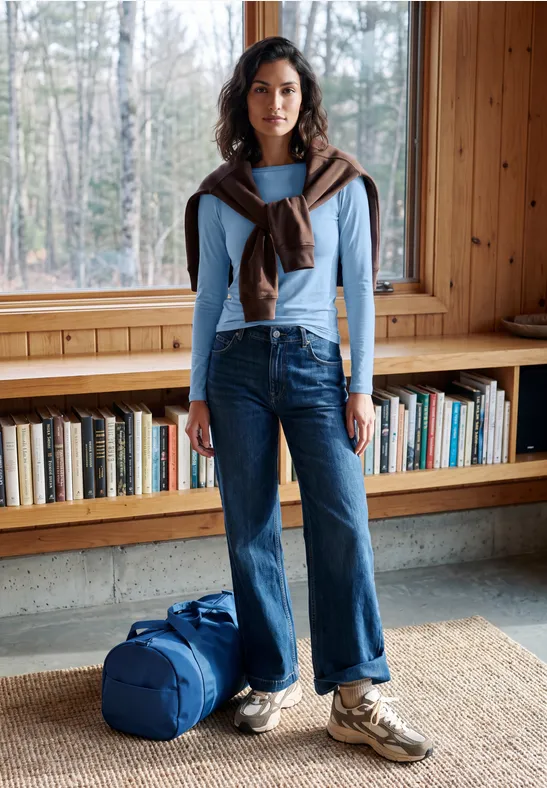 Frau in hellem Pullover, Jeans und Sneakers mit Reisetasche vor Bücherregal in Holzzimmer.
