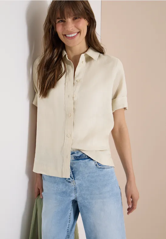 Eine Frau trägt beige Kurzarmbluse und helle Jeans, lehnt lässig an einer Wand und hält eine grüne Tasche.