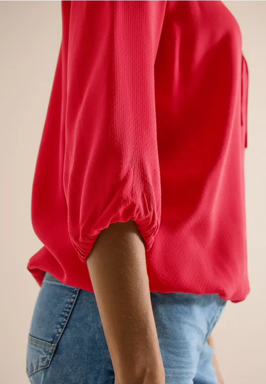 Seitliche Aufnahme einer Person in roter Bluse mit 3/4-Ärmeln und blauen Jeans.
