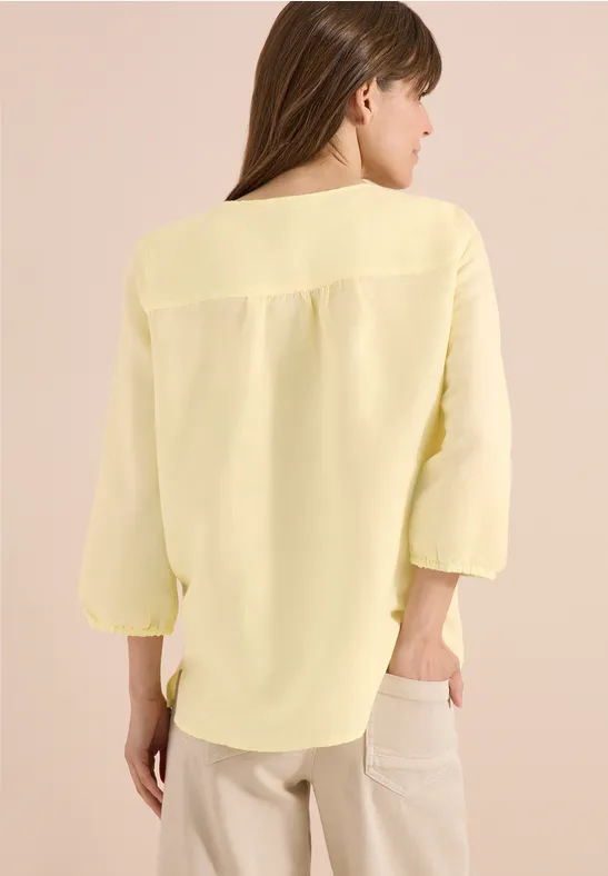 Femme de dos portant un chemisier jaune clair à manches 3/4 et un pantalon clair sur fond neutre.