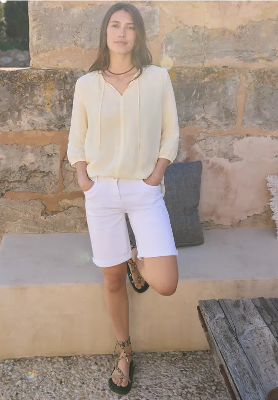 Personne en top clair et bermuda blanc appuyée décontractée contre un mur de pierre, un pied sur un banc.