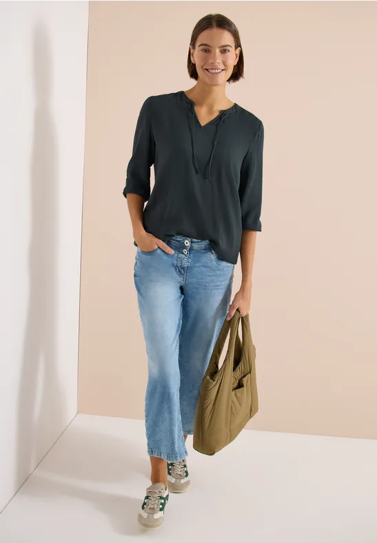 Frau steht vor hellem Hintergrund, trägt dunkles Oberteil, blaue Jeans, Sneaker und eine grüne Umhängetasche.