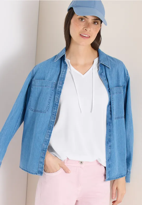 Person trägt hellblaue Jeansjacke, weißes T-Shirt, rosa Hose und hellblaue Mütze, mit Hand in Hosentasche.