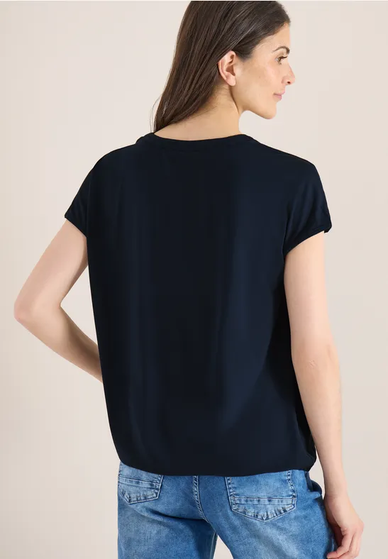 Frau von hinten in schwarzem Kurzarmshirt und blauen Jeans vor hellem Hintergrund.