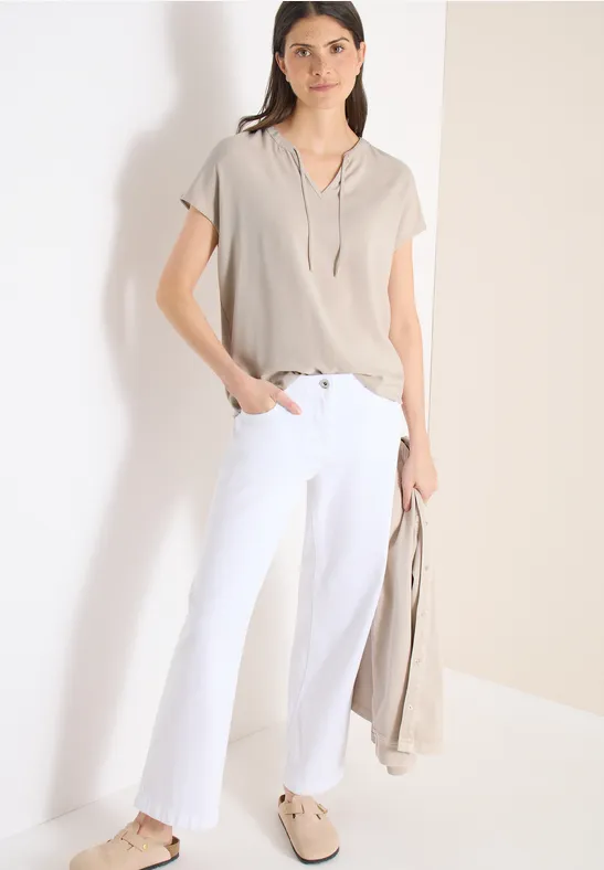Frau in beiger Bluse, weißen Hosen und beige Loafers, hält eine passende Jacke.