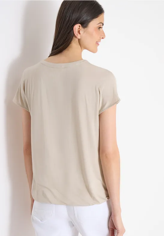 Person von hinten in beigem T-Shirt mit kurzen Ärmeln und weißer Hose vor hellem Hintergrund.