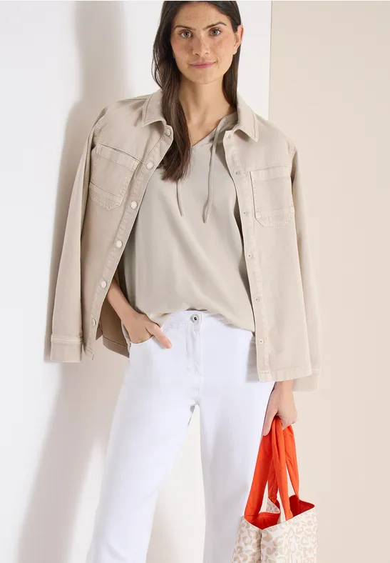 Eine Frau trägt einen beigen Blazer über den Schultern, ein helles Top, weiße Hose und hält eine orange Tasche.