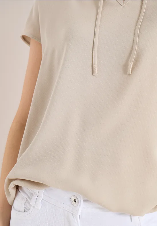 Beiges mouwelles Kapuzenshirt mit Kordeln und weißer Hose.