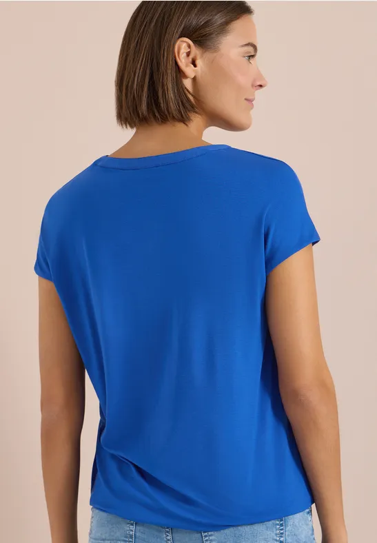 Eine Frau von hinten in einem schlichten, blauen T-Shirt vor neutralem Hintergrund.