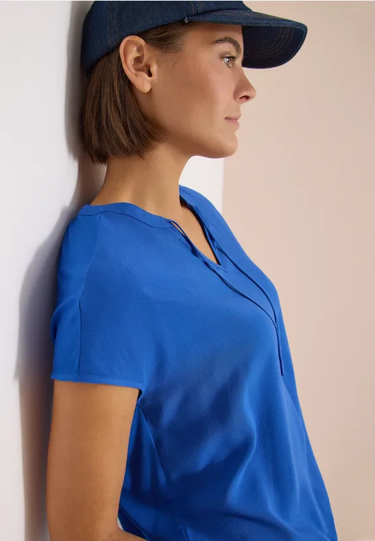 Frau mit blauem Kurzarmtop und dunkelblauem Basecap lehnt an heller Wand.