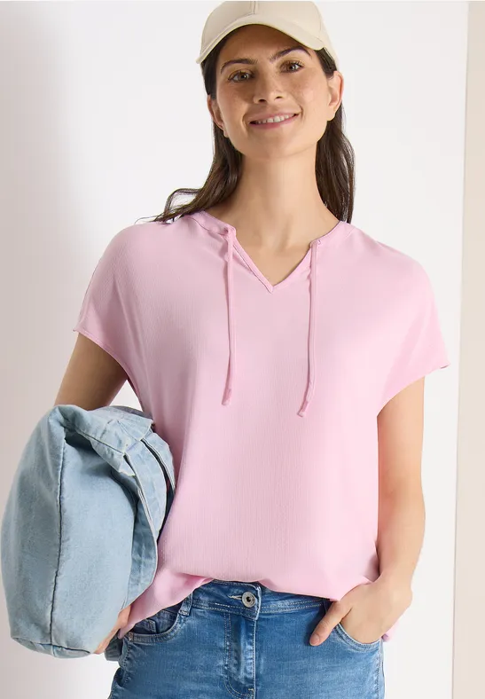Femme en t-shirt à capuche rose et jean, tenant un gilet bleu clair, coiffée d'une casquette blanche.