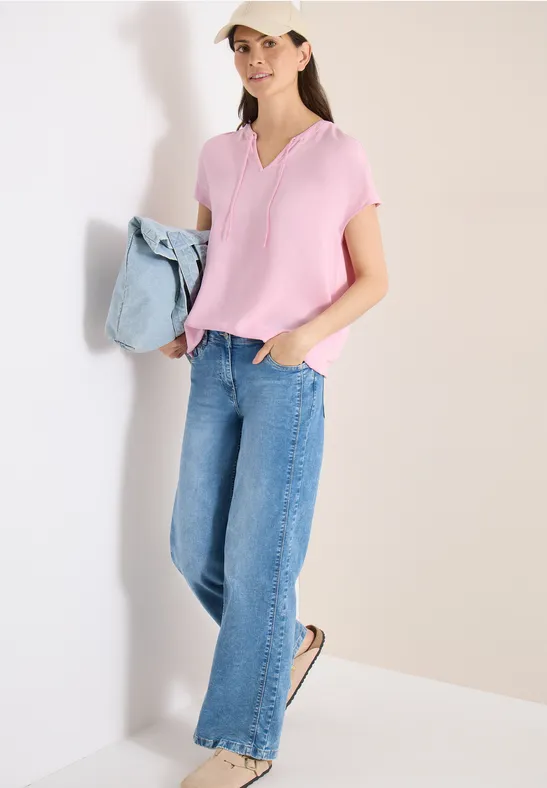 Femme en top rose et jean large, baskets blanches, tenue casual avec un pull bleu clair à la main.