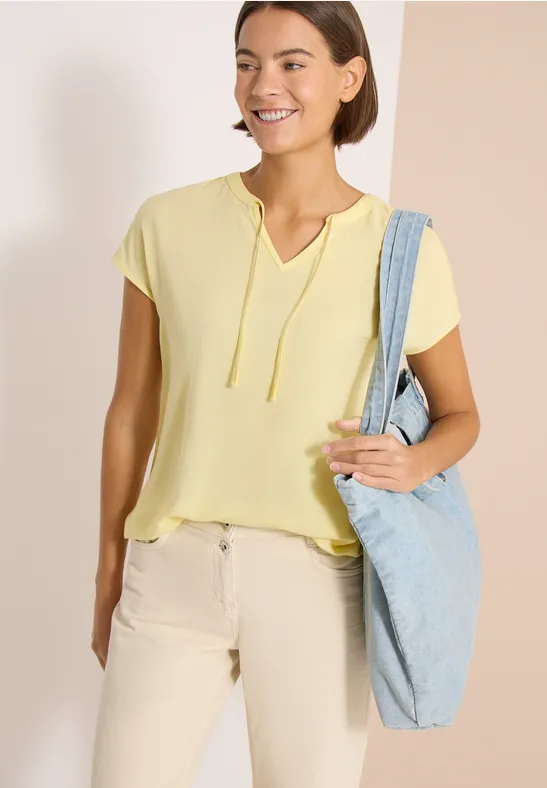 Frau mit gelbem Kurzarm-Shirt trägt eine hellblaue Schultertasche, neutraler Hintergrund.