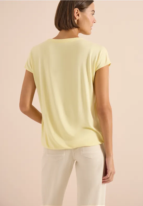 Person von hinten in hellgelbem Oberteil und heller Hose vor beigem Hintergrund.